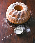 Bundt cake with icing sugar on cake rack beside sieve with icing sugar