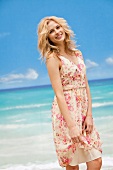 Beautiful blonde woman wearing floral print summer dress standing and smiling on beach