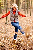 Cheerful blonde woman wearing gray jacket and jeans playing with autumn leaves in forest