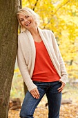 Portrait of happy blonde woman wearing white sweater leaning against tree, smiling
