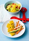 Potato salad in bowl and polenta slices with carrots, cucumber and radish on plate