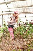 Cheerful blonde woman with long wavy hair wearing hat looking at roses in greenhouse