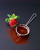 Strawberry half coated with love beads and chocolate besides tea strainer and cocoa powder
