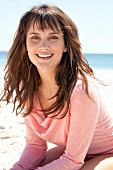 Portrait of pretty woman with brown hair wearing pink shirt standing on beach, smiling