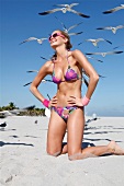 Frau in buntem Bikini kniet am Strand, hat Spaß, Möwen am Himmel
