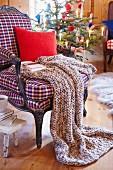 Gestrickte Decke mit Kissen und Buch auf Sessel, im Hintergrund Weihnachtsbaum