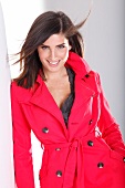 Portrait of attractive dark haired woman wearing red trench coat leaning against wall