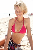 Pretty blonde woman in red bikini bottom and colourful cloth sitting on sand, smiling