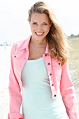 blonde Frau am Strand in rosa Kurzjacke, lacht, hat Spaß