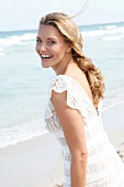 Portrait of happy blonde woman wearing white dress standing on beach, smiling