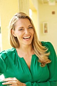 blonde Frau in grüner Bluse lacht in die Kamera, freundlicher Blick