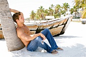 Contemplative blonde man sitting on beach and leaning against palm tree in front of boat