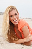 Portrait of happy blonde woman with long hair wearing orange top lying on beach, smiling