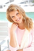 Portrait of pretty blonde woman wearing pink sweater sitting on chair at poolside, smiling