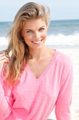 Portrait of pretty blonde woman wearing pink sweater sitting on beach, smiling