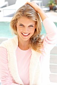 Portrait of pretty blonde woman wearing pink sweater sitting on chair at poolside, smiling