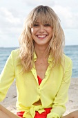 Portrait of cheerful blonde woman wearing yellow shirt sitting on beach, smiling