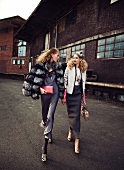 Two women wearing fur jacket talking while walking on street with old houses in background
