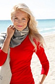 Portrait of pretty blonde woman wearing scarf over red sweater standing on beach, smiling