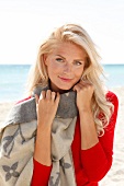 Portrait of pretty blonde woman wearing scarf over red sweater standing on beach, smiling