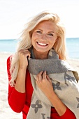blonde Frau im roten Pulli lächelt in Kamera, am Strand