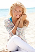 blonde Frau im hellen Pulli lächelt in Kamera, sitzt am Strand