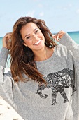 Pretty dark haired woman in gray sweatshirt with animal motif standing on beach, smiling