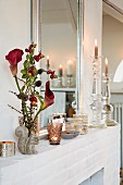A vase of calla lilies and sprigs of berries, tealights and candle holders on a mantlepiece in front of a mirror