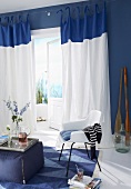 A blue and white living room with a fabric stool and a chair in a seating area in front of a balcony door with floor-length curtains