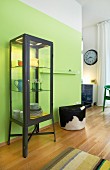 An industrial-style glass cabinet with LED lighting against a lime-green dining room wall next to a fur-covered pouffe