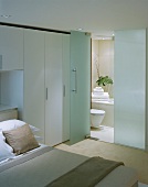Modern, white bedroom with open glass door and view into a bathroom ensuite