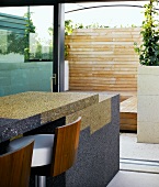 Bar aus Granitsteinen mit eleganten Barhockern vor offener Terrassentür und Blick auf Holztrennwand