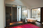 Freistehende Badewanne vor Terrassenfenster neben grosszügigem verglastem Duschraum und mit pinkfarbenem Streifenteppich