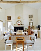 White-painted, carved wooden chairs at a wooden table with white sofas in the background in front of a fireplace