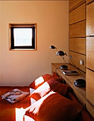 Orange Kissen auf Bett unter kleinem Fenster und Nachttischlampen auf Holzablage