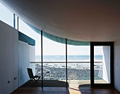 Empty living room with view of neighbouring beach through glass facade under concrete roof curving up as if floating on one side