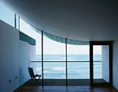 Empty room with view of terrace with chair and sea through glass facade under concrete sail curving up on one side