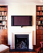 weiße Wand mit Flachbildfernseher über offenem Kamin zwischen traditionellen Bücherregalen