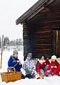 Fröhliche Familie beim Würstchen grillen am Lagerfeuer vor rustikaler Berghütte in verschneiter Winterlandschaft