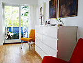Hell erleuchtetes Wohnzimmer mit rotem Sessel und gelbem Stuhl neben weißer Kommode