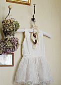 Weisses Babykleid mit Schühchen an einem Wandhaken neben getrockneten Blumensträussen