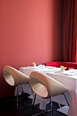 Set tables for two with white and deep pink designer chairs against brick-red wall