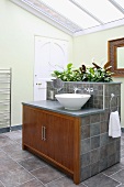 Modern bathroom with glass roof and grey tiles; wooden doors on central washstand island with integrated planter