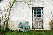Zwei Holzstühle an der Hauswand neben einer verwitterten Tür