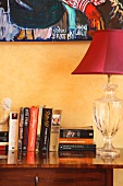 Books next to red lamp with decorative glass base on antique chest of drawers