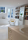 Teilweise sichtbare Badewanne mit eingebautem Regal und moderner Kunststoffstuhl vor Bücherwand im Schlafzimmer