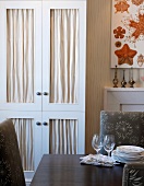 Dining table with crockery in front of elegant fitted cupboard with fabric panels in doors