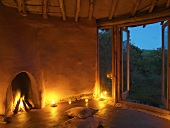 Burning open fire, candlelight and floor cushions in front of open terrace door in simple clay house