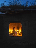 Clay house with green roof at dusk - view into interior lit by fire in fireplace and candlelight