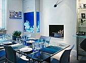 Zeitgenössisch maritimes Ambiente - gedeckter Glastisch vor blau leuchtendem Schrank-Aquarium und Feuerschale im Kamin